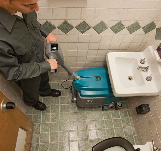 A Tennant T1B Battery Walk-Behind Scrubber cleaning a restroom. A Tennant T1B Battery Walk-Behind Scrubber cleaning a restroom.